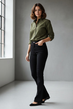 A woman stands indoors near a window, wearing a green button-up shirt styled with SPANX Pull On Flare Jean in Obsidian Rinse and black shoes. The background is minimal and gray.