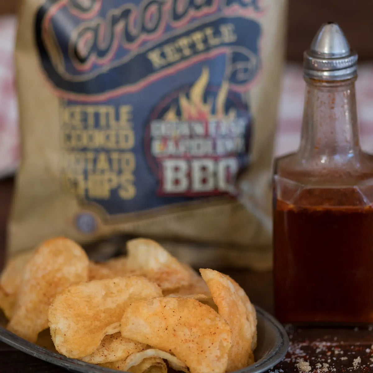 A bag of CAROLINA KETTLE - DOWN EAST BBQ CHIPS from 1 IN 6 SNACKS, featuring a brown and orange design with BBQ flames and bold text, delivers the authentic taste of Eastern North Carolina BBQ sauce accented by a hint of smoky charcoal flavor—making it a true standout among snack foods.