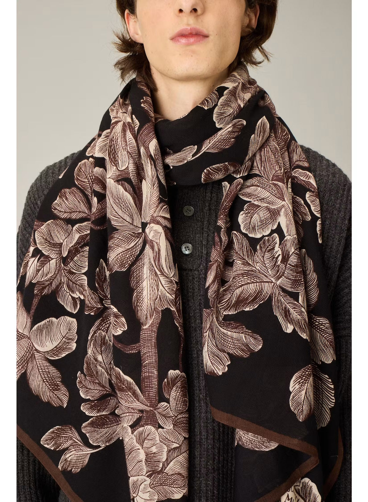 A person stands facing forward, dressed in a cream-colored, loose-fitting outfit featuring wide-legged pants and a long-sleeved top. They have paired their ensemble with black pointed shoes and the INOUI EDITIONS - ARBORIS WOOL SCARF IN BLACK by INOUI, which drapes elegantly over one arm. Made in India from 100% wool, the scarf adds a touch of luxurious comfort against the plain light background.