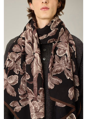 A person stands facing forward, dressed in a cream-colored, loose-fitting outfit featuring wide-legged pants and a long-sleeved top. They have paired their ensemble with black pointed shoes and the INOUI EDITIONS - ARBORIS WOOL SCARF IN BLACK by INOUI, which drapes elegantly over one arm. Made in India from 100% wool, the scarf adds a touch of luxurious comfort against the plain light background.