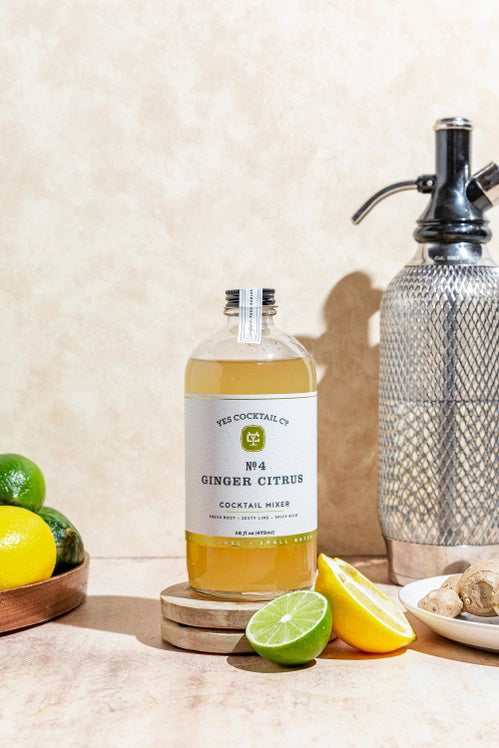 A clear glass bottle with a black cap containing Yes Cocktail Co.'s Ginger Citrus Cocktail Mixer is adorned with a white label featuring green accents. The text indicates it is crafted from fresh root, zesty lime, and has a spicy kick. The bottle holds 16 fl oz (473 ml).