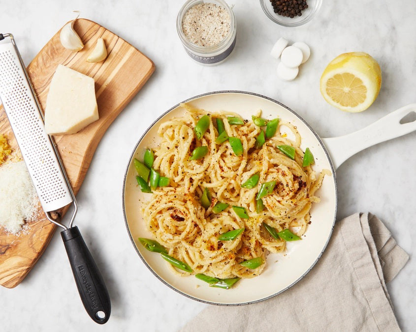 A small glass jar of JACOBSEN SALT CO. JACOBSEN SALT - INFUSED BLACK PEPPER SALT with a white lid rests on a light beige surface against a light gray background. The dark label with white text highlights this floral, spicy blend.