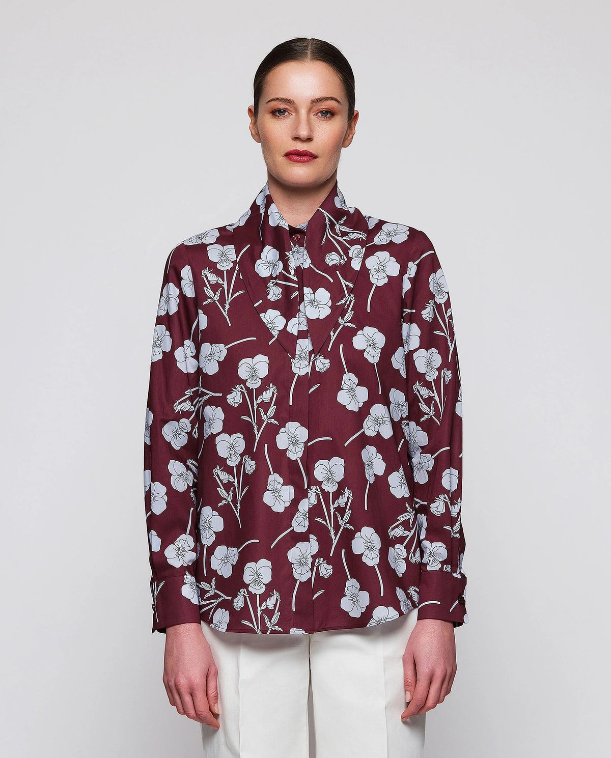 A woman stands by a window in a well-lit modern room, wearing the MIRTO 1956 Floral Print Blouse with Tie Neck in burgundy and high-waisted white pants, her hands casually tucked into her pockets.
