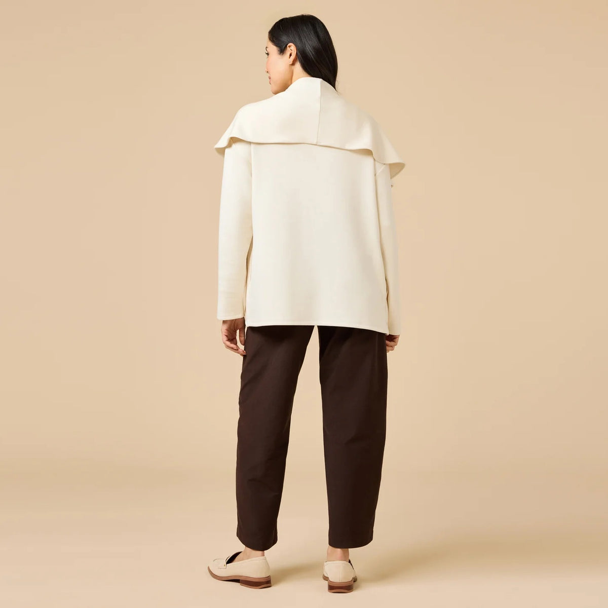 A woman wears an off-white SOFTIES - DREAMTECH EXAGGERATED COLLAR CARDIGAN with an exaggerated waterfall collar, cinched at the waist with a thin belt, and holds a matching handbag.