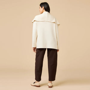 A woman wears an off-white SOFTIES - DREAMTECH EXAGGERATED COLLAR CARDIGAN with an exaggerated waterfall collar, cinched at the waist with a thin belt, and holds a matching handbag.