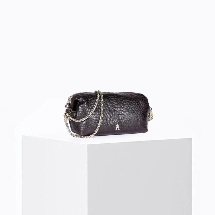 The CRAIE STUDIO - NUAGE BUBBLE POURPRE MINI BAG, a compact and textured black lambskin accessory featuring silver hardware and an eye-catching purple patterned strap, is showcased on a white pedestal against a plain background.