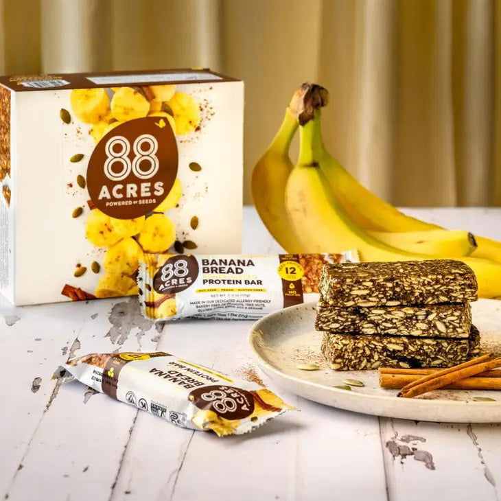 A protein bar wrapper labeled &quot;88 ACRES - BANANA BREAD HIGH PROTEIN BAR&quot; by 88 Acres. The wrapper highlights features such as &quot;Nut-Free,&quot; &quot;Vegan,&quot; &quot;Gluten-Free,&quot; 12 grams of protein, and &quot;No Palm Oil.&quot; The wrapper is set against a white wooden background.