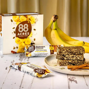A protein bar wrapper labeled "88 ACRES - BANANA BREAD HIGH PROTEIN BAR" by 88 Acres. The wrapper highlights features such as "Nut-Free," "Vegan," "Gluten-Free," 12 grams of protein, and "No Palm Oil." The wrapper is set against a white wooden background.