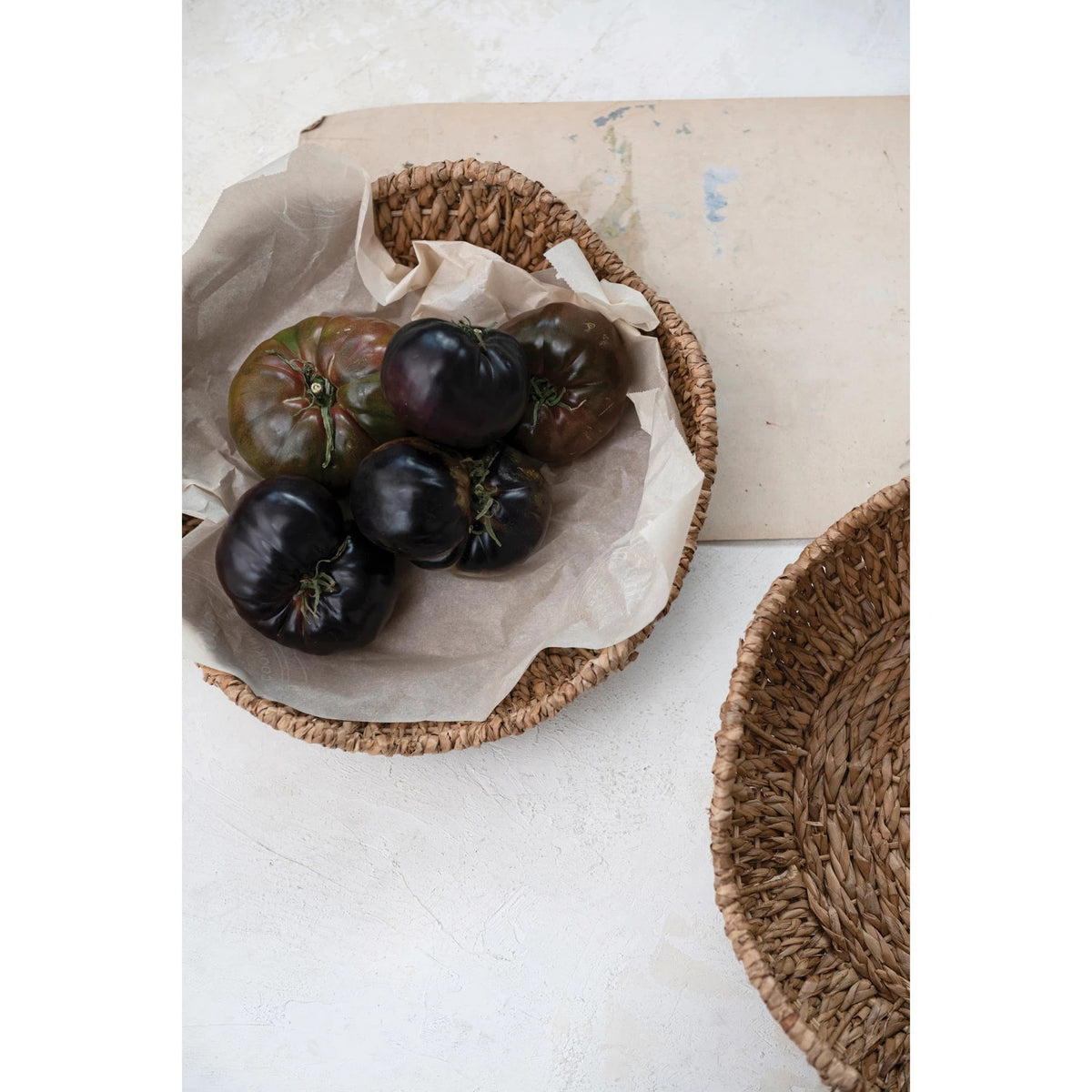 A wicker basket lined with parchment paper contains several dark purple tomatoes, positioned on a light-colored surface next to another empty &quot;BRAIDED BANKUAN BOWL WITH SCALLOPED EDGE (SMALL)&quot; by CREATIVE COOP and a piece of paper. The arrangement evokes a rustic or homey atmosphere.