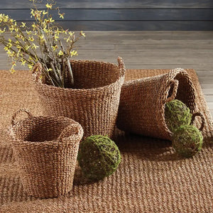 The NAPA HOME AND GARDEN Seagrass Tapered Basket with Handles and Cuff, Small, sits on a textured rug before a white paneled wall, alongside two other baskets, with part of a chair and a colorful painting in the background.