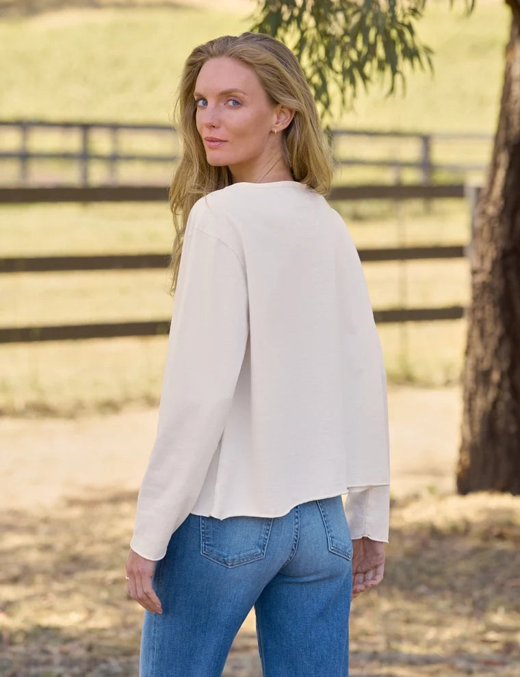 A woman with long blonde hair stands outdoors in front of a wooden fence, wearing the FRANK & EILEEN ELLA Long Sleeve Boatneck Tee in Vintage White and blue jeans, with trees and grassy fields in the background.