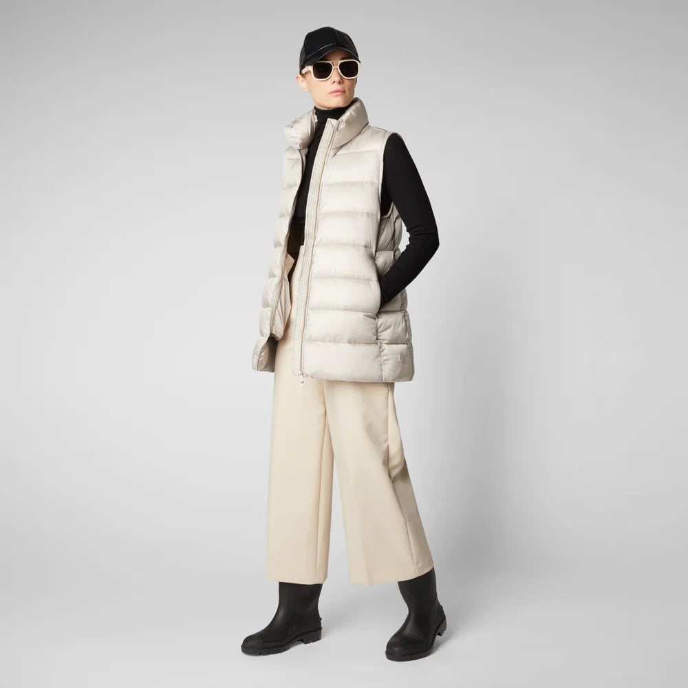 A person wearing a black cap, sunglasses, the SAVE THE DUCK - CORAL PUFFER VEST with animal-free down from SAVE THE DUCK over a black long-sleeve top, beige wide-legged pants, and black ankle boots stands against a plain background. The outfit appears to be styled for colder weather.