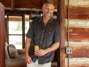 Close-up of a person wearing the CRIQUET Short Sleeve Corduroy Pearl Snap Shirt in Iron Horse, featuring pearl snap buttons and a chest pocket. Part of the person’s neck and chest is visible above the collar.
