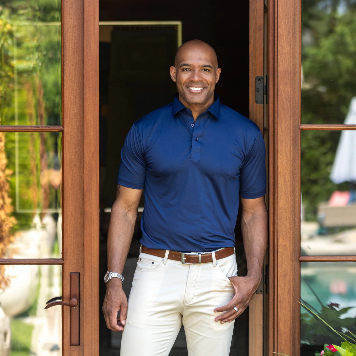 A man in the COLLARS AND CO FEATHERWEIGHT FLEX POLO IN NAVY and white pants stands by a light-paneled wall, smiling and holding the side of his performance golf shirt.