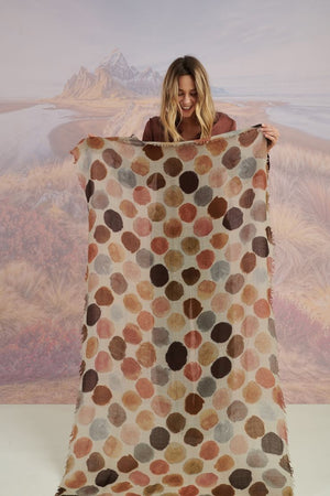 A woman with blonde hair displays the STORIATIPIC - BETTY WOOL SCARF, a large rectangular wool accessory with earth-toned polka dots, set against a scenic mountain landscape.