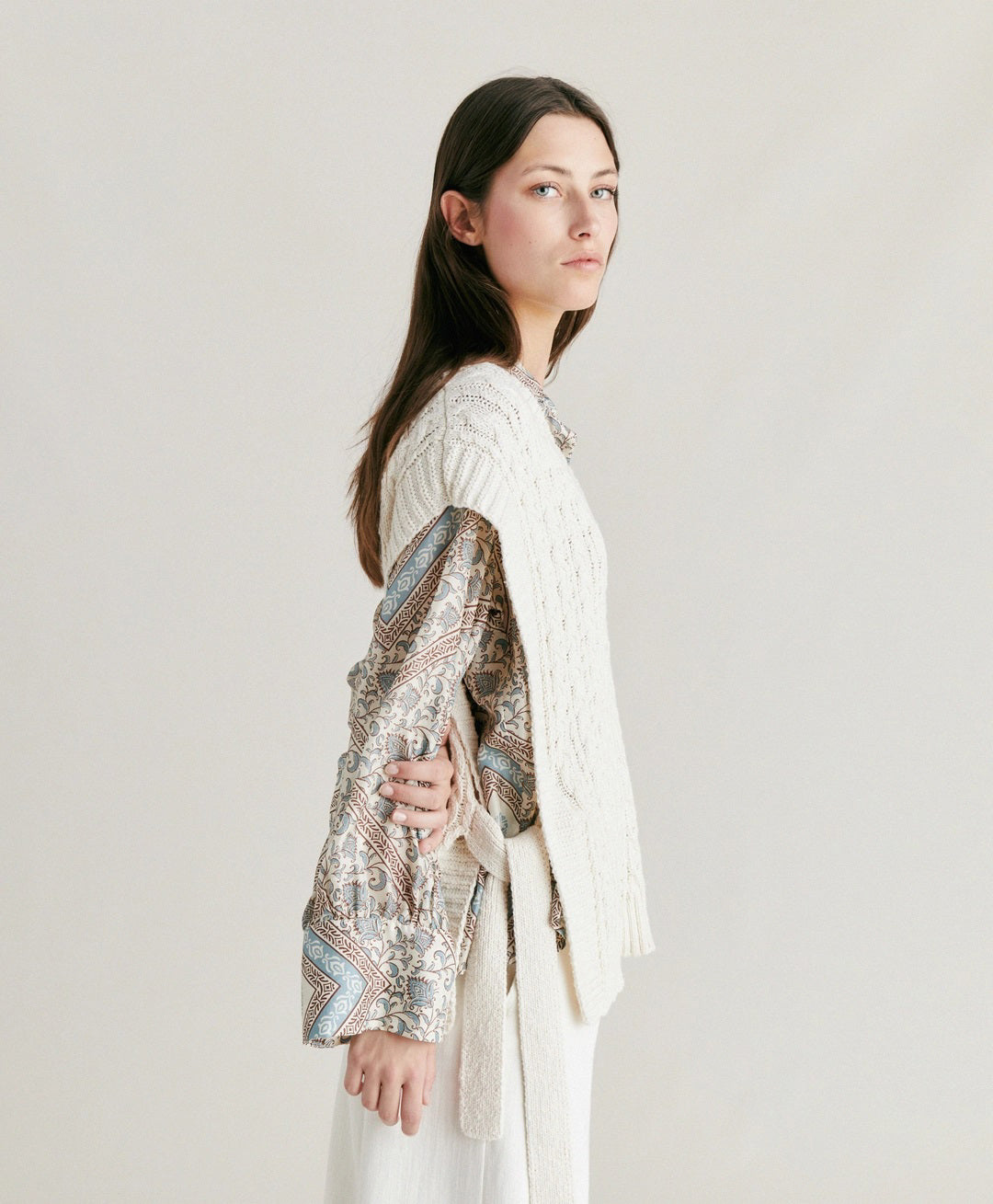 A woman with straight brown hair stands against a plain background. She is wearing the MOMONI - ARENE KNIT VEST from MOMONI, an oversized, cream-colored cable knit vest made from cotton yarn, over a patterned, long-sleeve button-up shirt and paired with white pants. Her hands are in her pockets, and she has a neutral expression.