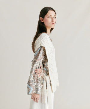 A woman with straight brown hair stands against a plain background. She is wearing the MOMONI - ARENE KNIT VEST from MOMONI, an oversized, cream-colored cable knit vest made from cotton yarn, over a patterned, long-sleeve button-up shirt and paired with white pants. Her hands are in her pockets, and she has a neutral expression.