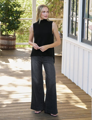 A person stands on a wooden deck, wearing the FRANK & EILEEN West Village NYC Wide Leg Trouser in Italian Dream Light Denim 1981 Wash paired with open-toe sandals. Only the lower half of their body is visible, set against a backdrop of greenery and a white structure, capturing effortless NYC style.
