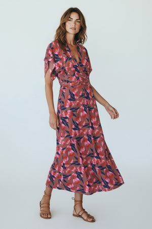 A woman stands indoors wearing the FRANCO FERRARI - JERSEY PRINT DRESS, a long, deep V-neck jersey dress with short sleeves and a red and blue botanical print. She pairs it with red shoes against draped white fabric and wooden frames.