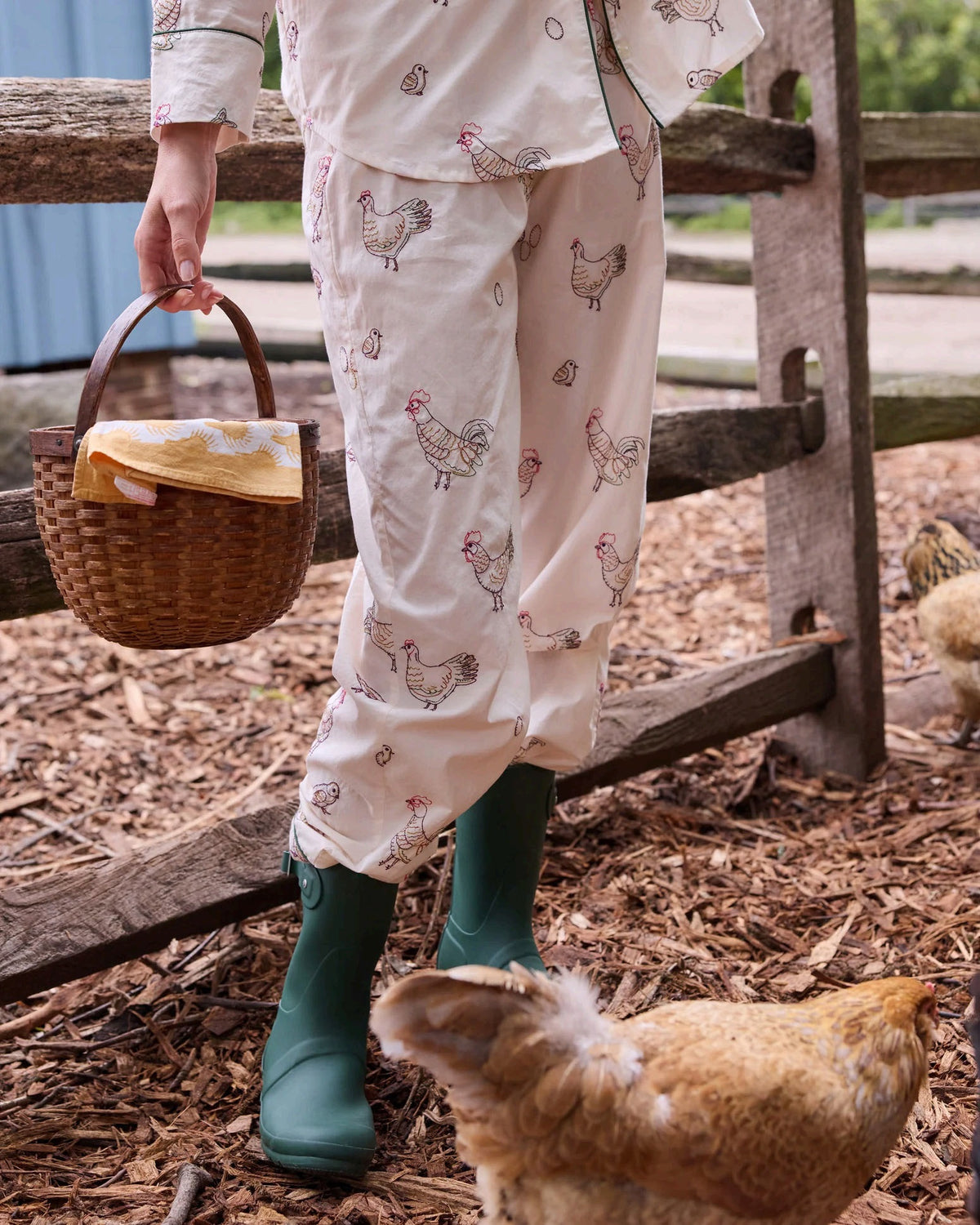 A woman dressed in the PRINTFRESH Embroidered Hens Long Pajama Set in Buff smiles as she stands by a wooden fence, holding a basket and watching chickens peck at the ground in a farmyard setting, with trees and a path visible behind her.