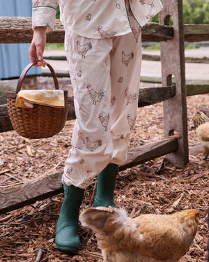 A woman dressed in the PRINTFRESH Embroidered Hens Long Pajama Set in Buff smiles as she stands by a wooden fence, holding a basket and watching chickens peck at the ground in a farmyard setting, with trees and a path visible behind her.