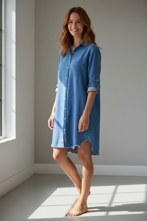 The woman stands outside in a FRANK & EILEEN Mary Classic Shirtdress in vintage stonewashed indigo, holding a beige bag and smiling slightly, with green bushes, a large planter, and a light-colored building facade in the background.