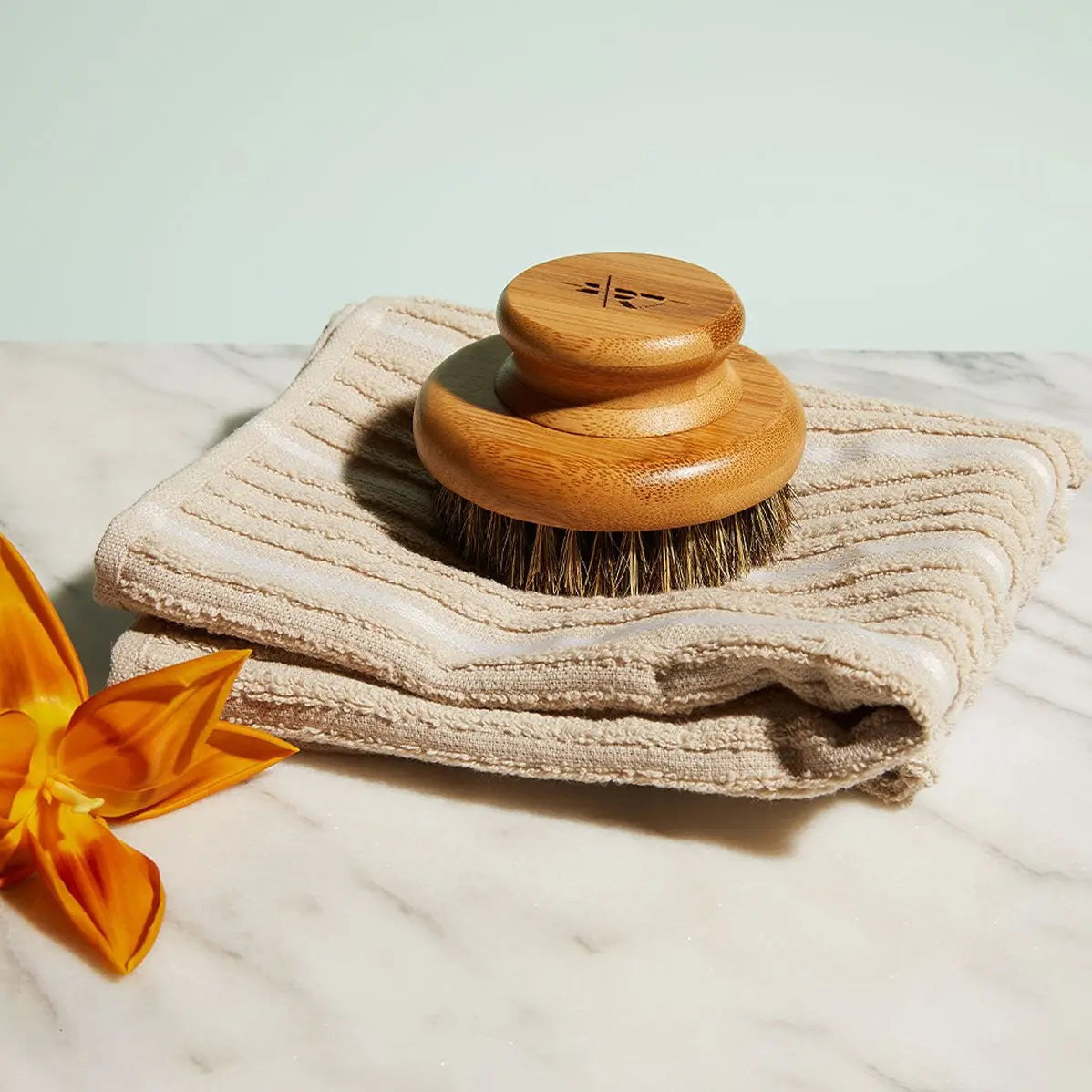The KATE MCLEOD - THE DRY BRUSH, featuring natural bristles, is displayed on a beige towel atop marble, accented with an orange flower—evoking a relaxing dry brushing and lymphatic drainage ritual.