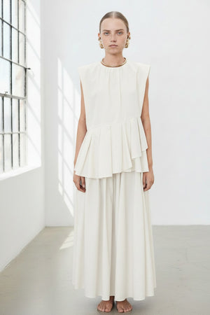 The individual is elegantly clad in the MEIMEIJ PLEATED ASYMETRICAL TOP, a sleeveless brown design with a unique peplum hem, paired with a matching long skirt. Gold hoop earrings and red flat shoes add flair against the plain white backdrop.