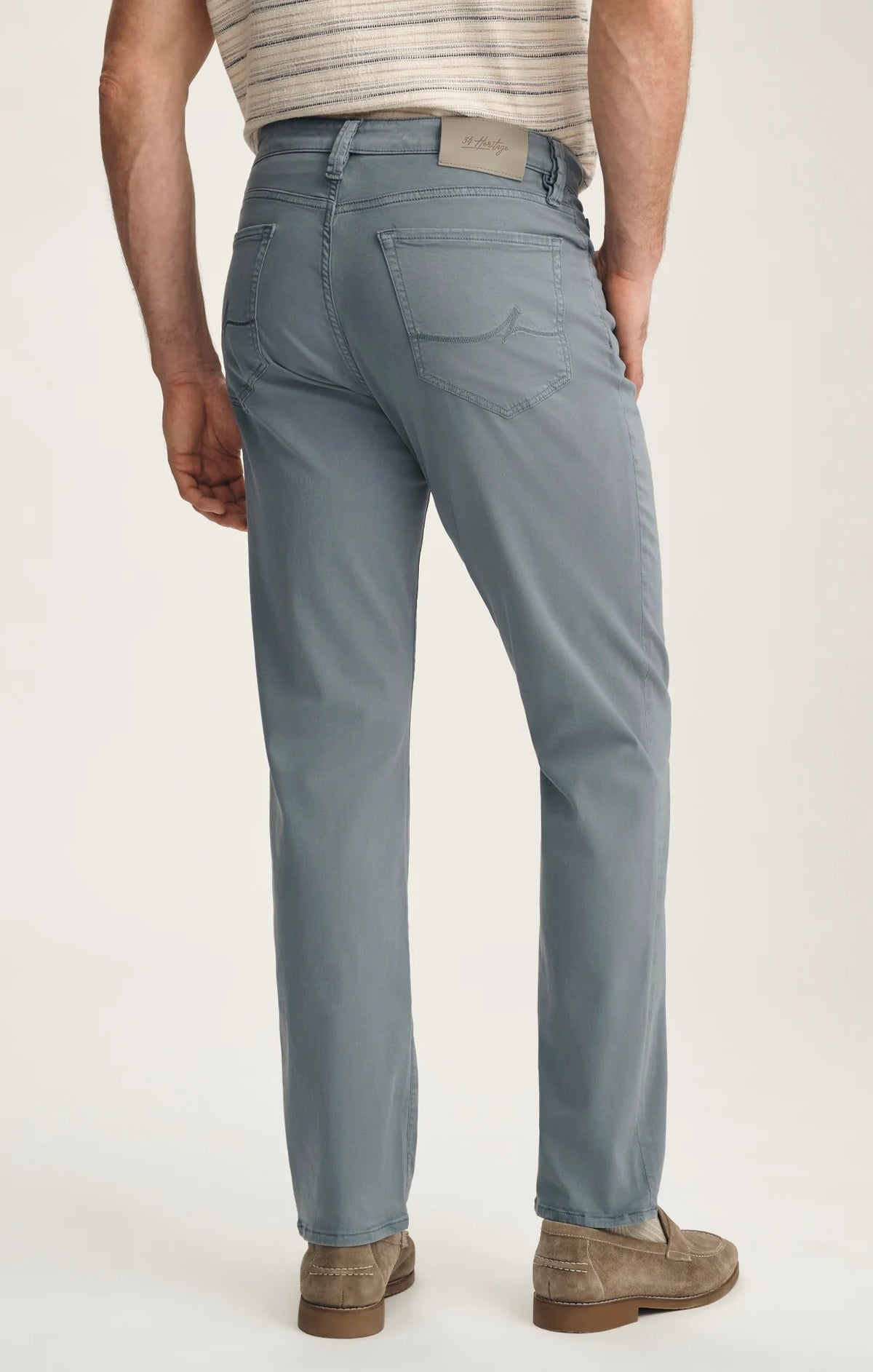 A man stands with his back to the camera against a plain light background, wearing a beige striped short-sleeve shirt, 34 HERITAGE Charisma Relaxed Straight Leg Pant in Flint Stone, brown suede shoes, and sunglasses.