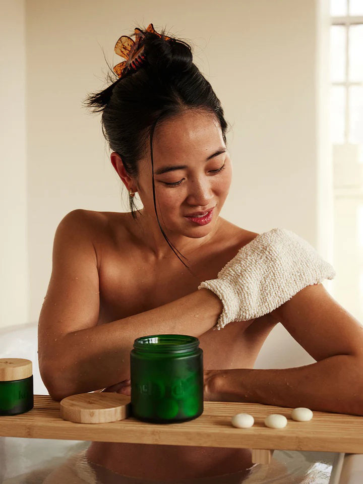 A hand showcases a textured beige KATE MCLEOD - THE BODY MITT against a light turquoise background, while another hand adjusts the cuff with its black tag and red stitching. Beneath, the white surface featuring gray veining creates an ideal exfoliating spa treatment ambiance.
