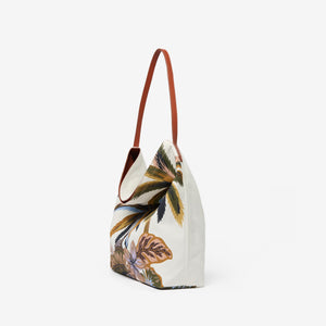 The INOUI EDITIONS - HAWAII CAMPUS CANVAS BAG highlights a tropical landscape in earthy tones with palm trees and foliage, offering a painterly vintage charm. Its slouchy design and single leather strap elegantly combine style and function against a plain white background.
