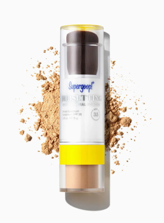 A container of SUPERGOOP 100% MINERAL POWDER SPF35 with built-in brush sits on a white surface, surrounded by scattered light brown powder. This SUPERGOOP powder sets makeup and controls shine.