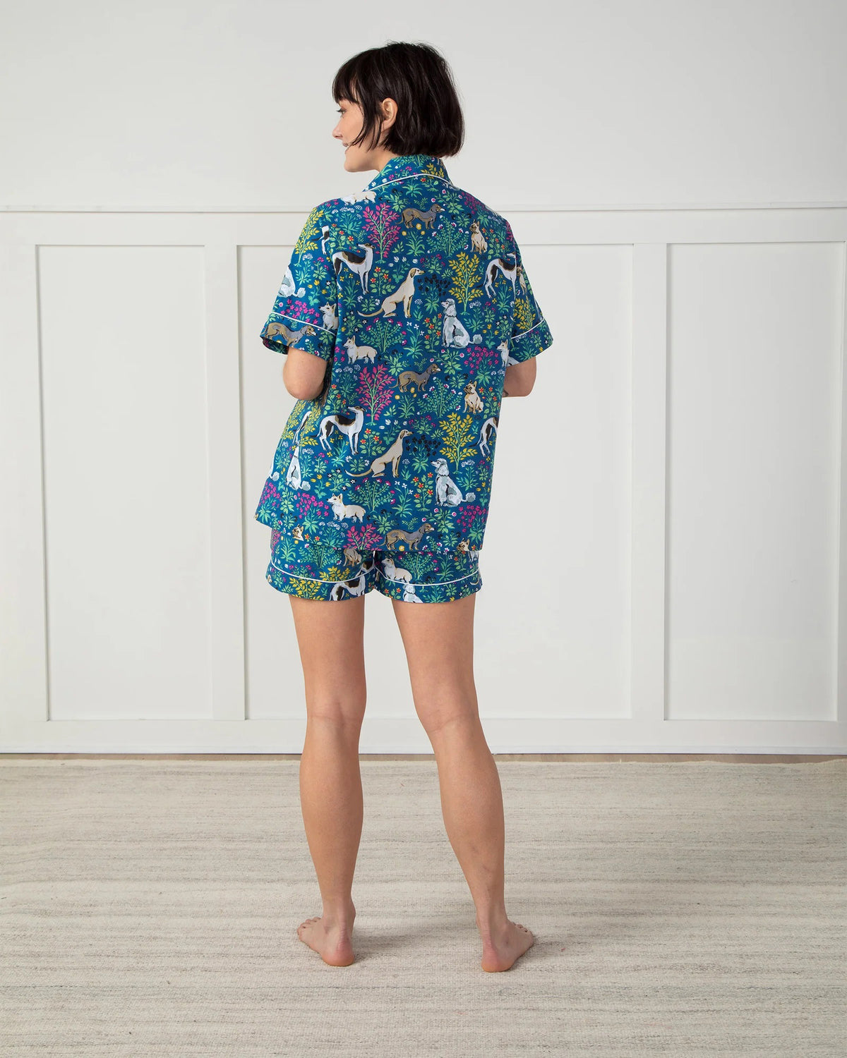 A person stands against a plain white wall wearing the stylish PRINTFRESH - MUST LOVE DOGS PAJAMA SHORT SET made of organic cotton. The set, adorned with colorful dog and floral illustrations on a blue background, features pockets where the person&#39;s hands rest.