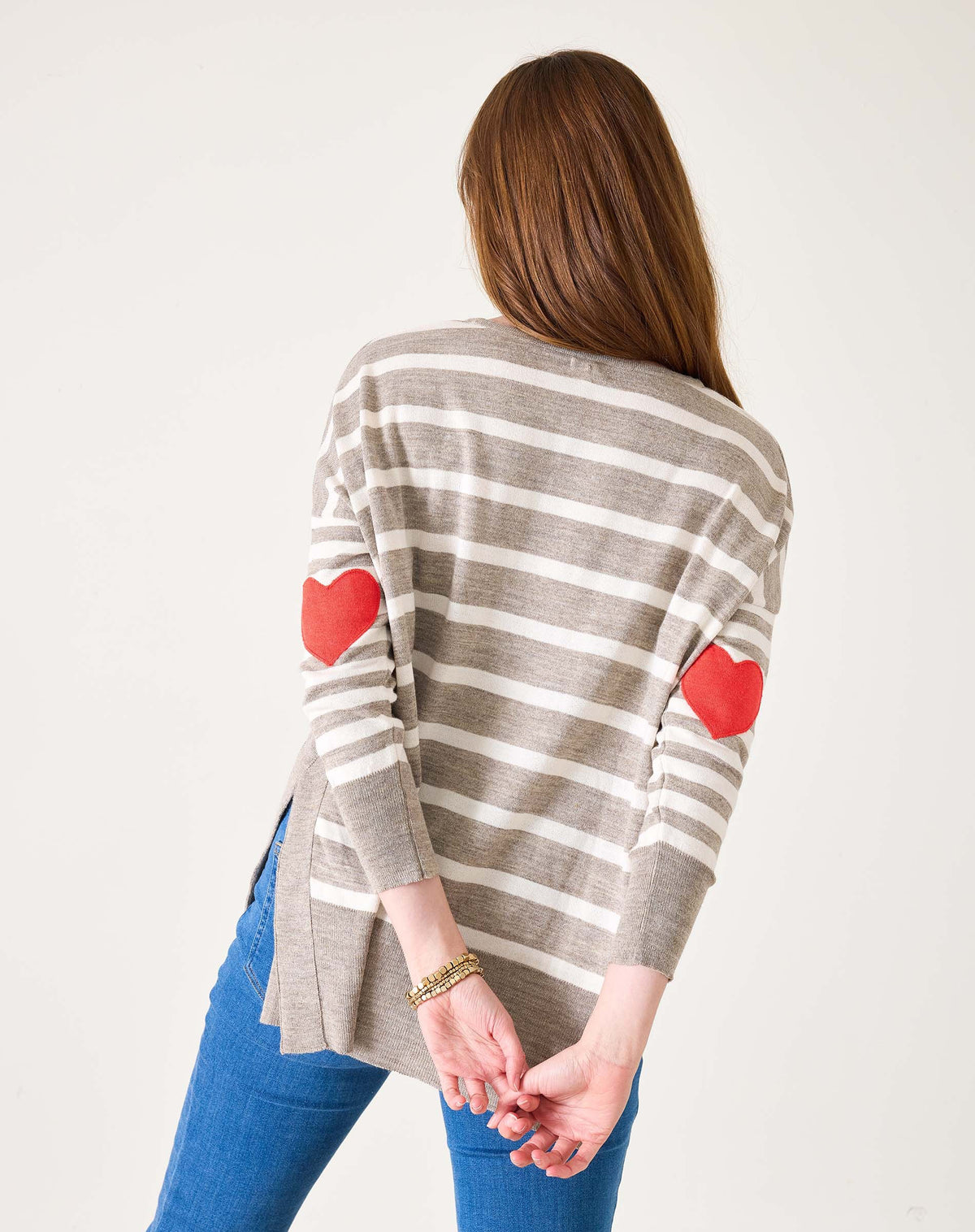 A woman with long brown hair in blue jeans models the MERSEA - AMOUR SWEATER IN DRIFTWOOD STRIPE by MERSEA, featuring gray and white stripes with red heart-shaped elbow patches. She stands sideways against a plain white background.