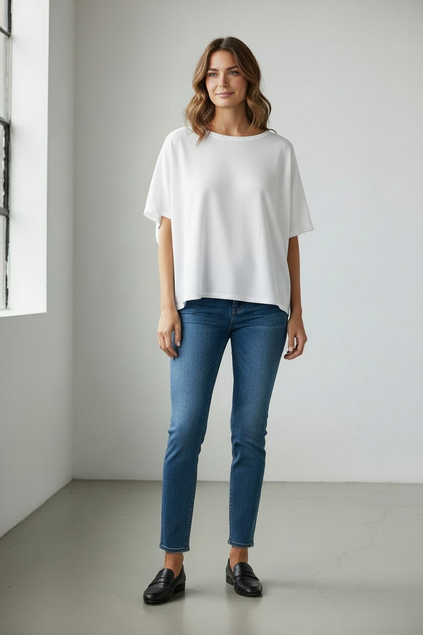 A woman with wavy brown hair stands near a window in a minimalist room, smiling softly with her hands by her sides. She is wearing the FRANK & EILEEN Olive Original Capelet in White—a loose, 100% cotton short-sleeve top—paired with blue jeans and black loafers.