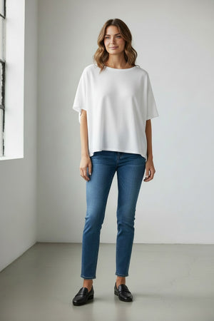 A woman with wavy brown hair stands near a window in a minimalist room, smiling softly with her hands by her sides. She is wearing the FRANK & EILEEN Olive Original Capelet in White—a loose, 100% cotton short-sleeve top—paired with blue jeans and black loafers.