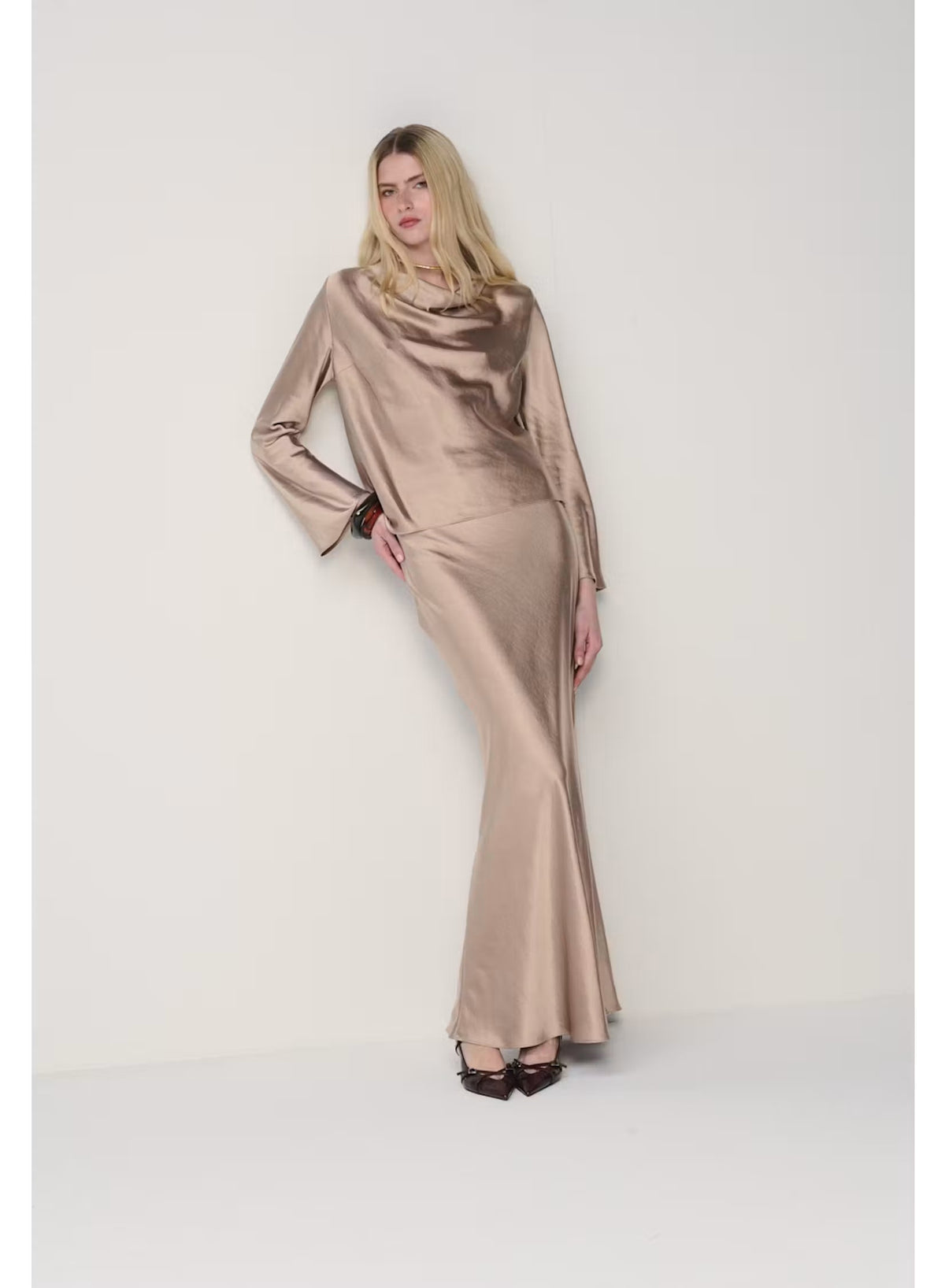 A person wearing the MILVA MI - LONG SATIN SKIRT IN SAND by MILVA, paired with pointed black shoes and standing against a plain, light background.