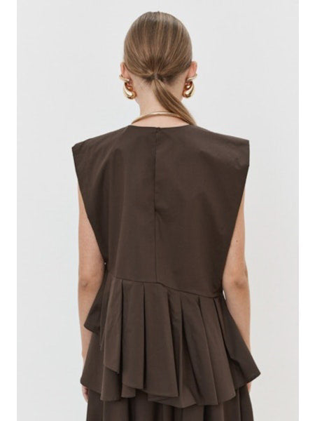 The individual is elegantly clad in the MEIMEIJ PLEATED ASYMETRICAL TOP, a sleeveless brown design with a unique peplum hem, paired with a matching long skirt. Gold hoop earrings and red flat shoes add flair against the plain white backdrop.