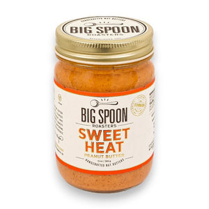 A spoonful of BIG SPOON ROASTERS - SWEET HEAT PEANUT BUTTER against a plain white background. The peanut butter appears creamy with visible small chunks of peanuts, giving it a slightly rough texture. The spoon is metallic and reflects some light, highlighting the spicy essence of this popular condiment from BIG SPOON ROASTERS.
