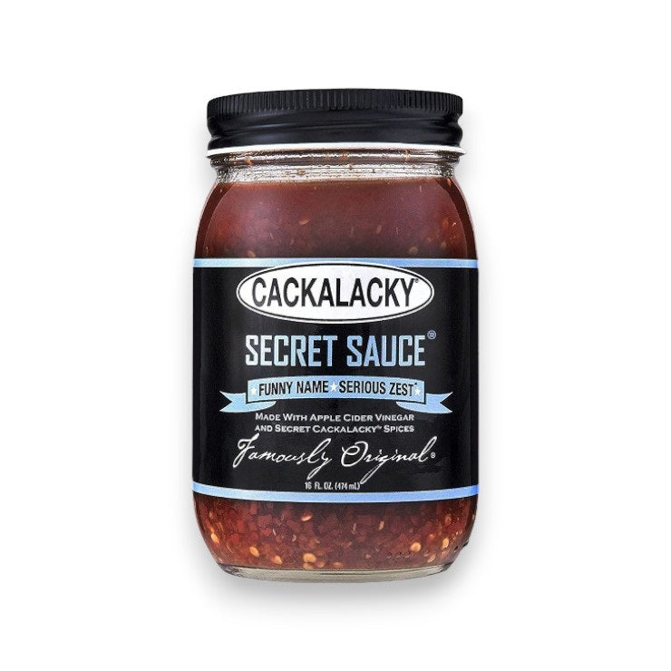 A 16 fl oz (473 ml) jar of CACKALACKY - SECRET SAUCE, topped with a black lid and adorned with a label that reads "Funny Name • Serious Zest," highlights its artisanal apple cider vinegar and signature Cackalacky spices. Visible seasoning within the jar attests to its authenticity as a true Carolina vinegar sauce.