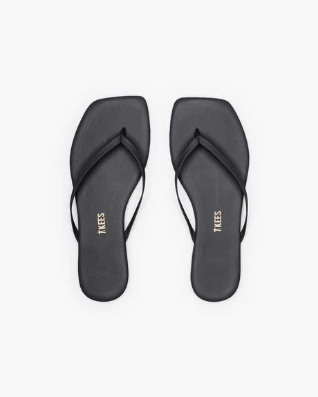 The TKEES INC SQUARE TOE LILY BLACK sandals feature a black leather upper with thin straps and square toes, shown from above on a white background. &quot;TKEES&quot; is printed in gold on the insoles, emphasizing their modern design.