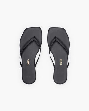 The TKEES INC SQUARE TOE LILY BLACK sandals feature a black leather upper with thin straps and square toes, shown from above on a white background. "TKEES" is printed in gold on the insoles, emphasizing their modern design.