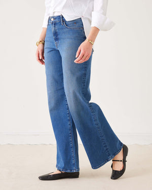 A person wearing the MERSEA Kate Wide Leg Jean in Baltic Wash, featuring four-way-stretch technology, paired with a white long-sleeve shirt with rolled cuffs, black flat shoes, and gold bracelets, stands on a light-colored floor against a plain white background.