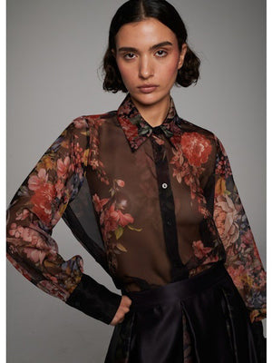 A woman with wavy dark hair models the MILVA MI - FLORAL VOILE BLOUSE by MILVA—a semi-sheer, long-sleeve top with a dark floral print and black collar—paired with a high-waisted skirt, standing before a plain gray background.