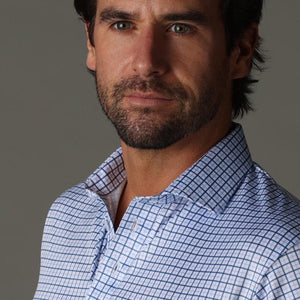 A man with dark hair and a beard wears the COLLARS AND CO - SEMI SPREAD COLLAR POLO IN BLUE TATTERSAL under a light brown sweater, standing against a gray background and looking to his right with a serious expression.