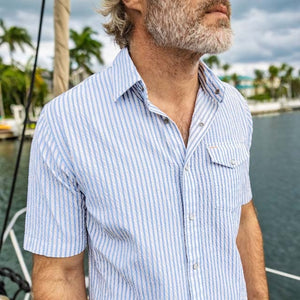 A smiling man with short gray hair and a beard wears the CRIQUET Short Sleeve Performance Seersucker Pearl Snap Shirt in Navy, featuring blue and white vertical stripes, paired with light pants against a plain white background.
