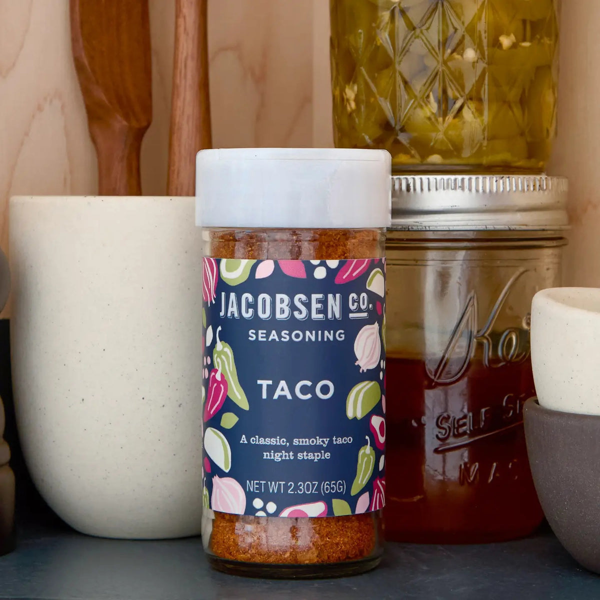 A jar of JACOBSEN SALT - TACO SEASONING from JACOBSEN SALT CO. rests on a kitchen shelf, alongside a white container, wooden utensils, a glass jar with a yellow lid, and a mason jar filled with honey.