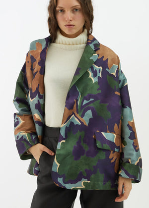 A person with long hair is sporting the ALYKI LABORATORIO PRINTED SATIN JACKET, featuring a green, purple, and brown floral pattern, over a white turtleneck sweater. With one hand in the pocket of their black pants, they perfectly blend an elegant color outfit against a plain white background.