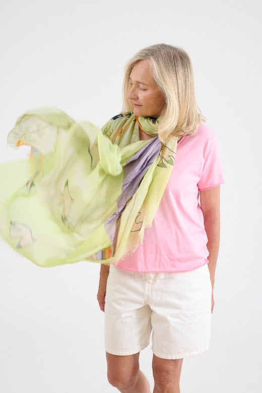 A woman with shoulder-length blonde hair wears a light pink shirt and the STORIATIPIC - GERTRUD SCARF IN ANISE, a colorful modal and silk accessory featuring green, yellow, and purple tones. She stands against a plain light background, looking directly at the camera.