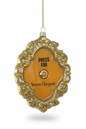 A 3.5-inch oval-shaped Cody Foster PRESS FOR FRENCH CHAMPAGNE ORNAMENT in a gold frame, featuring a center button, small inlaid jewels, and text that reads “PRESS FOR Veuve Clicquot.”.
